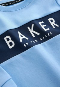 Világoskék pulóver sötétkék csíkkal, amelyen vastag fehér betűkkel szerepel a "BAKER" szó, alatta pedig a "by TED BAKER" felirat. Simább anyagtextúra.