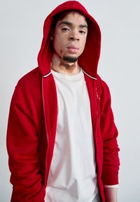 Sweat à capuche rouge à fermeture éclair avec une capuche doublée, présentant des accents blancs et une poche frontale, porté sur un t-shirt blanc uni. Texture de tissu lisse.