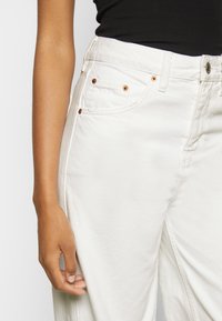 Calças de denim brancas com cintura alta, apresentando rebites de cobre nos bolsos, detalhe na costura lateral e uma superfície ligeiramente texturizada.