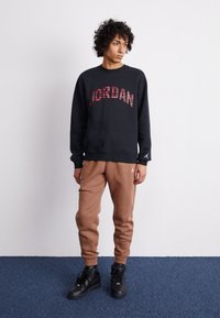 Black sweatshirt with "JORDAN" in plaid lettering, paired with brown sweatpants. Black sneakers complete the outfit. Casual style and fit.