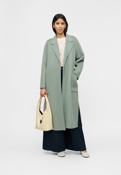 Γυναίκα που στέκεται με ένα μακρύ παλτό σε χρώμα φασκόμηλο, λευκή πλεκτή μπλούζα, φαρδιά σκούρα μπλε παντελόνια, μπεζ τσάντα και ανοιχτά παπούτσια slip-on σε λευκό φόντο.