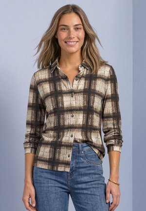 Une femme souriante porte une chemise boutonnée à carreaux beiges et bruns, aux manches retroussées, avec un jean bleu, sur un fond bleu pâle.
