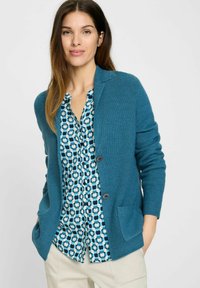 Türkiser gestrickter Cardigan mit ribbelner Textur, ausgestattet mit einem Schalkragen, zwei Taschen und Knopfverschluss, über einer gemusterten Bluse getragen.