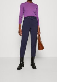 Ljuslila tröja, figursydd design, i kombination med mörkblå byxor med bälte. Svarta ankelstövlar och en brun väska. Släta tygtexturer.