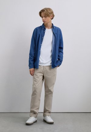 Teen boy with blond hair wearing blue button-up shirt over white t-shirt, beige pants, and white sneakers standing against plain wall.