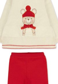Maglione crema con design di un orso, cappello rosso con pom-pom e pantaloni rossi. Il maglione presenta bordi a costine e un accento a fiocco. Texture complessiva morbida.