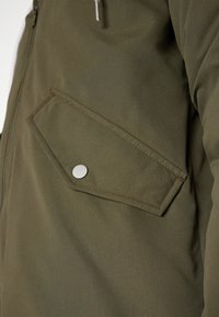 Bolsillo de chaqueta verde oliva con solapa inclinada y botón de presión plateado, que presenta costuras visibles y detalle de cremallera lateral.