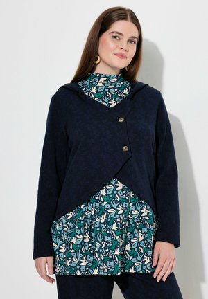 Marineblauer Kapuzencardigan aus strukturiertem Stoff, mit einem Überwurfdesign und zwei Knöpfen, über einem gemusterten teal- und weißen Oberteil getragen.