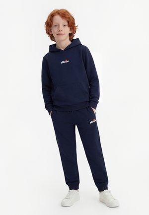 Dziecko z rudymi włosami, ubrane w granatową bluzę z kapturem ellesse i pasujące spodnie dresowe oraz białe trampki, stoi z rękami w kieszeniach.