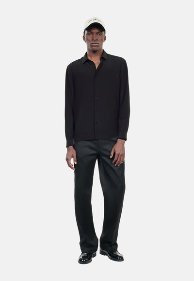 Sort langærmet skjorte med knapper, parret med sorte bukser. Tan kasket med logo og blanke sorte sko fuldender outfittet.