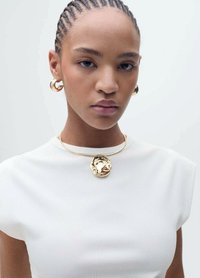 Colier cu pandantiv circular din aur și cercei hoop din aur purtați de un model cu păr texturat, îmbrăcat într-o bluză albă ajustată.