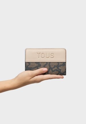 Mano sosteniendo una cartera TOUS de color beige y negro con un diseño estampado y el nombre de la marca en relieve, sobre un fondo claro liso.