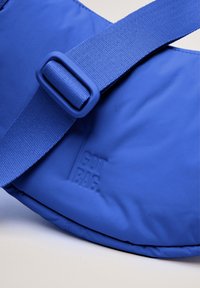 Borsa di stoffa blu con una larga tracolla dotata di fibbia, logo "GOT BAG" inciso e superficie liscia. Design complessivo elegante.