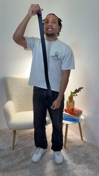 Čierny látkový opasok držený vertikálne, so striebornou prackou, kombinovaný so svetlým tričkom a tmavými džínami. Neutrálne pozadie, neformálne prostredie.