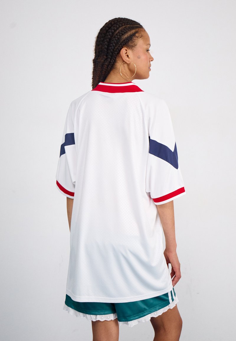 Weißes Sporttrikot mit roten und marineblauen Akzenten, kurzen Ärmeln und einem gerippten Kragen. Kombiniert mit grünen Shorts und Spitzenbesatz am Saum.