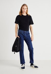 Camiseta de algodón negra, jeans de mezclilla azul oscuro con costuras en contraste, sosteniendo una mochila negra con un logotipo, usando zapatillas marinas con suelas blancas.