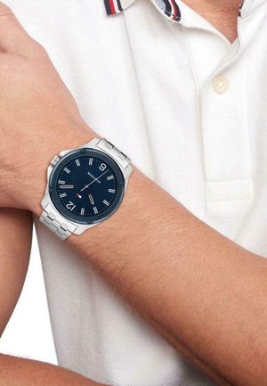Montre en acier inoxydable avec un cadran bleu marine, des index argentés et un design de bracelet décontracté. Portée sur un poignet avec une chemise polo blanche.