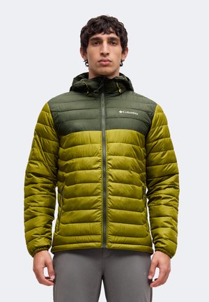 Joven con una chaqueta acolchada Columbia verde y oliva con capucha, de pie y mirando hacia adelante con un fondo liso.