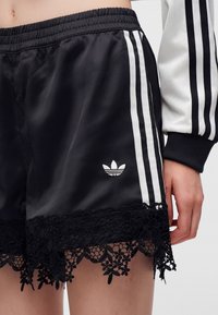 Short noir Adidas avec des bandes blanches et une bordure en dentelle, porté avec une manche de veste Adidas blanche et noire assortie visible.