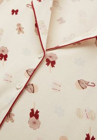 Beżowy materiał w świąteczny wzór z piernikami, babeczkami, lizakami i kokardami, wykończony czerwonymi lamówkami i guzikami.