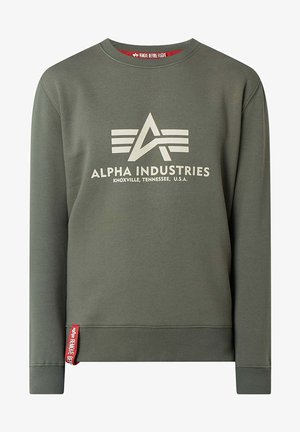 Oliivroheline pusakas, mis on valmistatud puuvilla segukangast, suur valge "Alpha Industries" logo ja punane silt allääres.