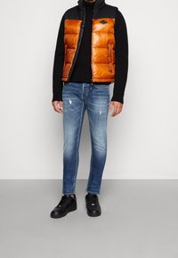 Fodrad orange väst med svarta detaljer, svart tröja, blå jeans med slitna detaljer och svarta sneakers. Avslappnad, lager-på-lager-outfit.