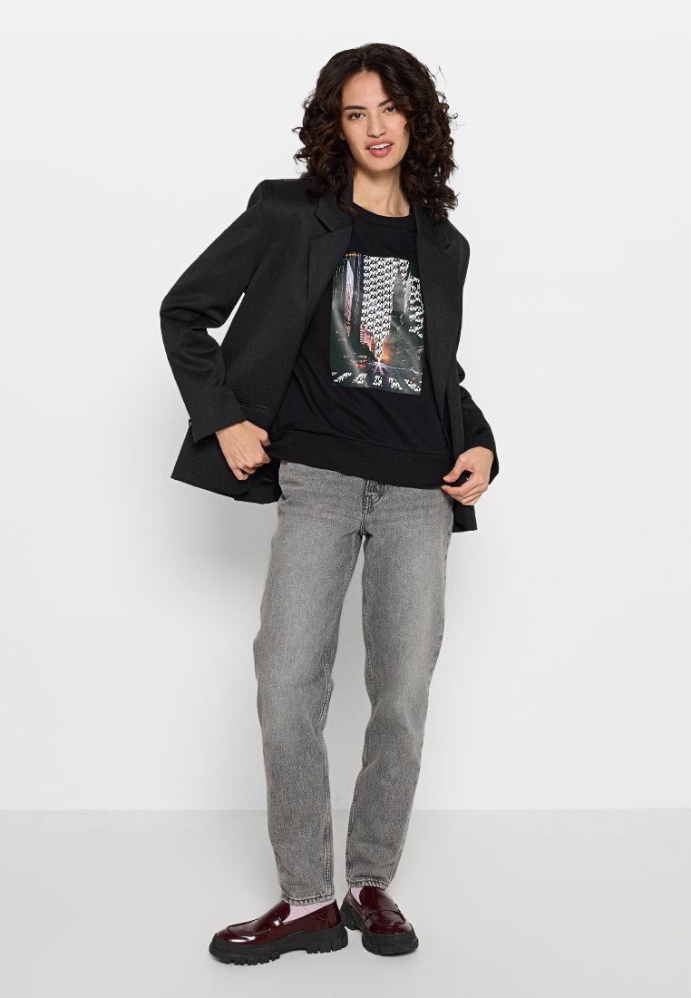 Blazer negro sobre una camiseta negra con un gráfico, combinado con jeans grises. El calzado es brillante, de color rojo oscuro. Corte recto con un ajuste relajado.