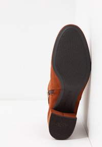 Botte en daim marron avec semelle antidérapante noire et détail de fermeture éclair, vue depuis le dessous sur un fond blanc en angle.