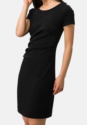 Femme portant une robe noire ajustée à manches courtes, longueur genou, debout devant un fond clair uni.