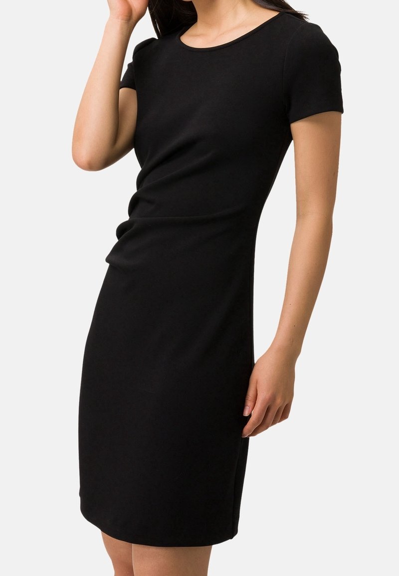 Femme portant une robe noire ajustée à manches courtes, longueur genou, debout devant un fond clair uni.