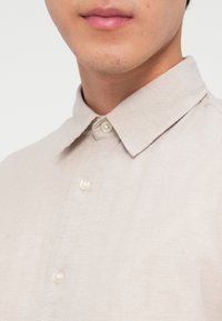 Κοντινό πλάνο του κάτω μέρους του προσώπου και του λαιμού ενός άνδρα που φοράει ένα ανοιχτό μπεζ πουκάμισο με κλασικό γιακά και κουμπιά.