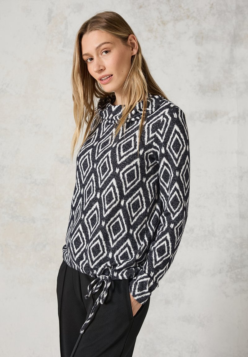 Svart och vit polotröja med diamantmönster, cowl-neck, långa ärmar och dragsko i nederkant. Matchas med svarta byxor.