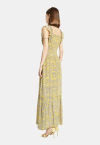 Abito maxi in stampa floreale gialla, corpino aderente con spalline fru-fru, gonna svasata, tessuto leggero. Indossato con sandali gialli con tacco.