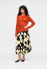 Κόκκινο πλεκτό πουλόβερ με στρογγυλή λαιμόκοψη και αφηρημένο σχέδιο, συνδυασμένο με μαύρη φούστα με πιέτες που φέρει κίτρινες και ροζ οργανικές μορφές. Μαύρες γόβες.