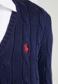 Cardigan en maille torsadée navy avec un logo de joueur de polo brodé en rouge sur une chemise à col blanc.