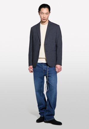Blazer texturé gris foncé avec deux boutons, pull en tricot crème en dessous, jeans larges bleus et bottes noires. Détail en chaîne sur les jeans.