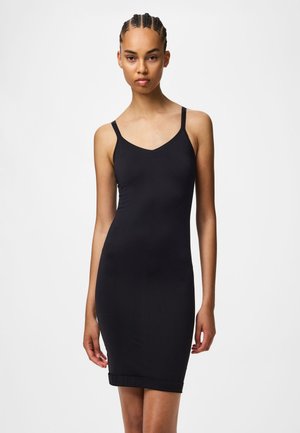 Žena s vrkočmi, oblečená v priliehavých čiernych šatách bez rukávov s tenkými ramienkami, stojaca pred jednoduchým bielym pozadím.