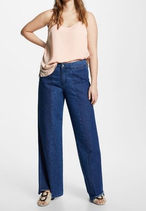 Femme portant un débardeur rose poudré, un jean large bicolore bleu, et des sandales beiges, debout avec une main sur la hanche.