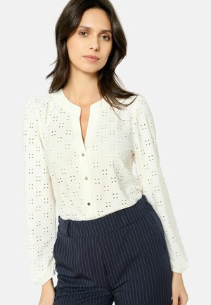 White long-sleeve blouse with eyelet pattern and button closure, paired with dark pinstriped trousers. Smooth texture, round neckline.