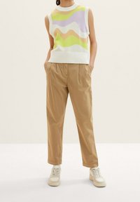 Bezramionny biały sweter z kolorowymi falistymi wzorami w zielonym, fioletowym i pomarańczowym; w połączeniu z beżowymi spodniami i jasnymi trampkami.