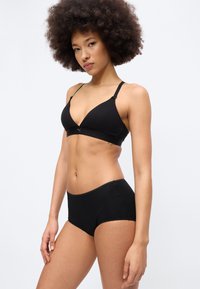 Γυναίκα με σγουρά μαλλιά που φοράει μαύρο αθλητικό σουτιέν και μαύρο εσώρουχο σε στυλ μποξεράκι, στέκεται μπροστά σε απλό λευκό φόντο.