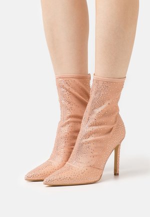 Nogi w cielistych, zaostrzonych botkach na obcasie do kostki, ozdobionych rozproszonymi małymi srebrnymi ćwiekami, na gładkiej powierzchni materiału.