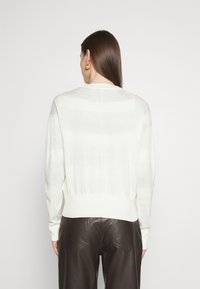 Svetle béžový svetr s dlouhými rukávy, žebrovaným lemem a jemným pruhovaným vzorem; nošený s tmavými koženými kalhotami.