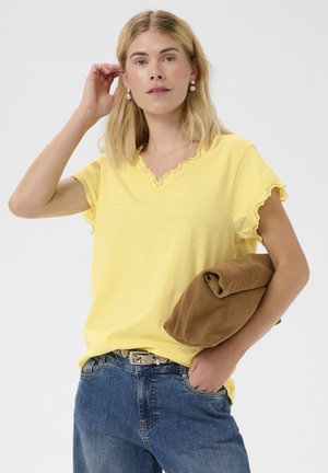 Blondynka w żółtej koszulce z dekoltem w serek i falowanym brzegiem, niebieskich jeansach z paskiem w wężowy wzór, perłowe kolczyki, trzyma złożoną brązową kurtkę.