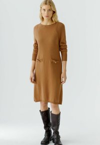 Braun gestricktes Kleid mit langen Ärmeln, geripptem Ausschnitt und zwei vorderen Taschen. Getragen mit kniehohen schwarzen Lederstiefeln.