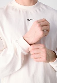 Koszula z długim rękawem w jasnym odcieniu kremowym z okrągłym dekoltem. Posiada czarne logo "HUGO" na piersi oraz prążkowane mankiety. Wykonana z miękkiej bawełny.