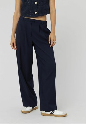 Pantalons larges bleu marine avec plis, en tissu léger et à la texture lisse. Associés à des baskets blanches avec des rayures noires.
