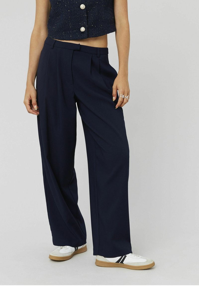 Pantalons larges bleu marine avec plis, en tissu léger et à la texture lisse. Associés à des baskets blanches avec des rayures noires.