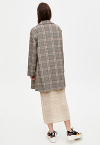 Rutig överrock i dämpade toner av grått, beige och ljusorange, kombinerad med en krämfärgad stickad kjol och svarta sneakers med färgaccenter.