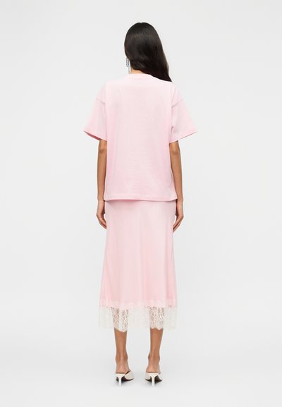 Ροζ υπερμεγέθη μπλουζάκι με κοντά μανίκια, σε συνδυασμό με μια ροζ φούστα μέσης μάκρους με λευκή δαντέλα στο τελείωμα, φορεμένα με ανοιχτόχρωμες γόβες.